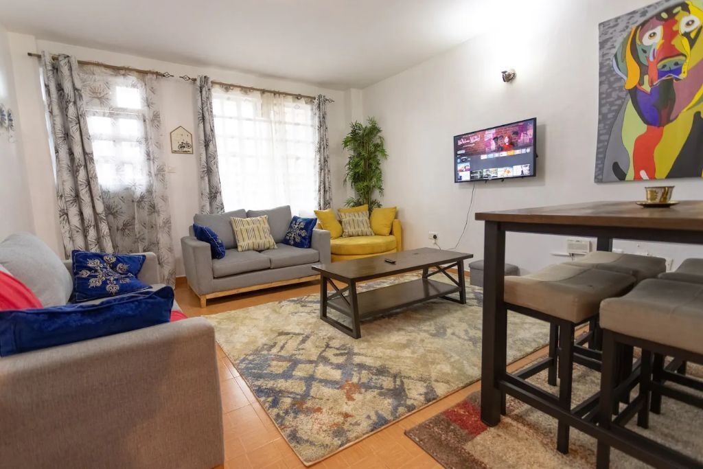 A living room with a grey sofa, yellow ottoman, and a coffee table. There is a TV mounted on the wall and a painting above it.