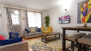 A living room with a grey sofa, yellow ottoman, and a coffee table. There is a TV mounted on the wall and a painting above it. - Cozy Family-sized 2 Bedroom Apartment with stable Wi-fi & conducive work station (Ruiru)