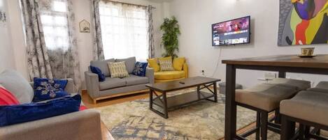 A living room with a grey sofa, yellow ottoman, and a coffee table. There is a TV mounted on the wall and a painting above it.