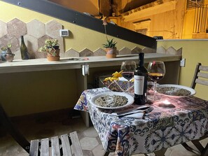 Uma mesa de jantar posta com pratos de comida, uma garrafa de vinho e copos, com um muro de azulejos e uma escada ao fundo.