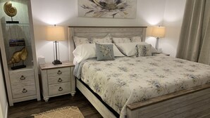A bedroom with a wooden bed, floral bedding, a nightstand with a lamp, a dresser with decorative items, and a rug. - Literally: 15 steps to the Heated Pool/Ground Floor Condo/King Bed/Free Parking (Clearwater)