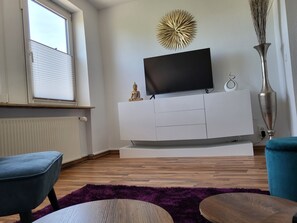 Una sala moderna con un televisor de pantalla plana sobre un mueble blanco, una estatua de Buda, un adorno de pared y un jarrón alto.
