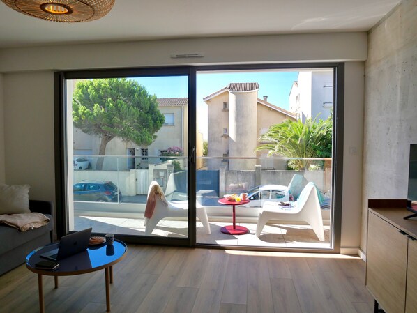 A modern living room with a glass sliding door leading to a balcony. Inside, there is a round table with a laptop, books, and a cup. The room has a sofa and a cabinet. - Luxury brand new appartment 40m away from a large sand beach (Mauguio)