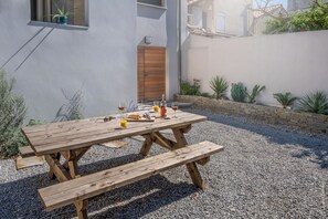 A wooden picnic table with benches, a bottle of wine, and a glass of orange juice on a gravel surface. - Luxury brand new appartment 40m away from a large sand beach (Mauguio)