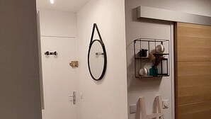 Käytävän lattia on puuta, siinä on valkoinen ovi, valkoinen patteri, valkoinen tikapuu ja seinässä musta pyöreä koriste.