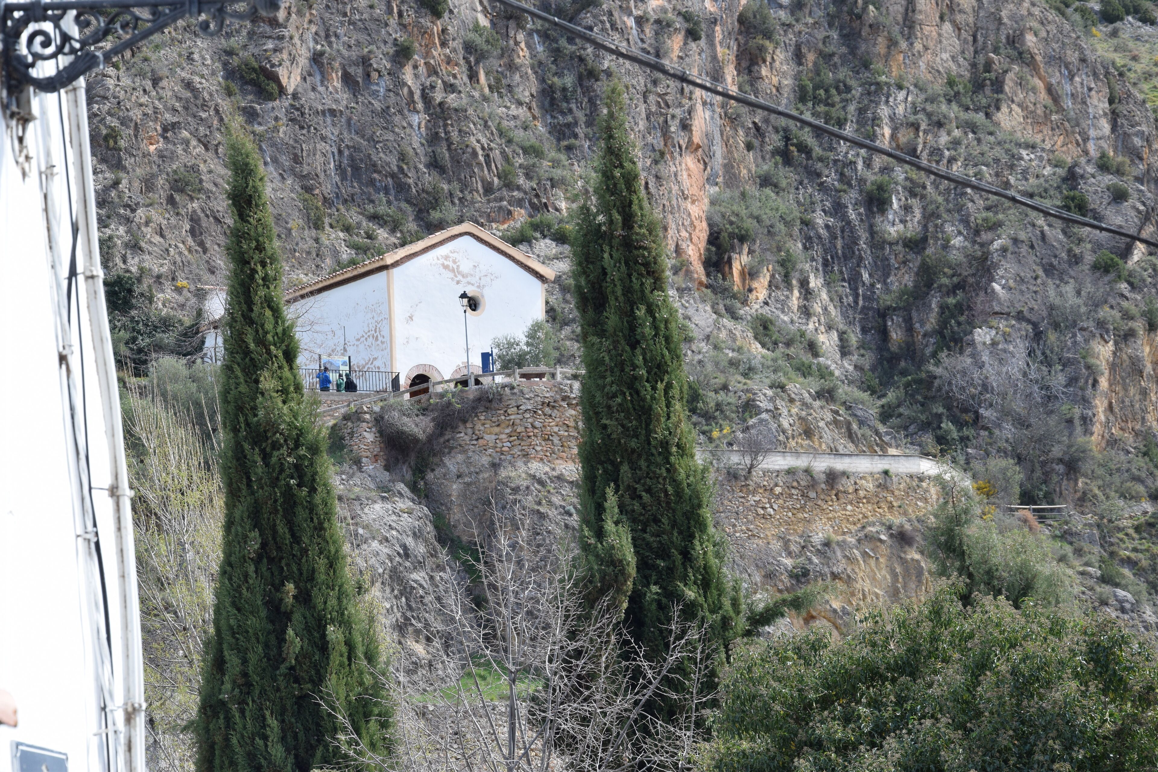 A building with a simple, white facade and a flat roof, situated on a rocky hillside with sparse vegetation.