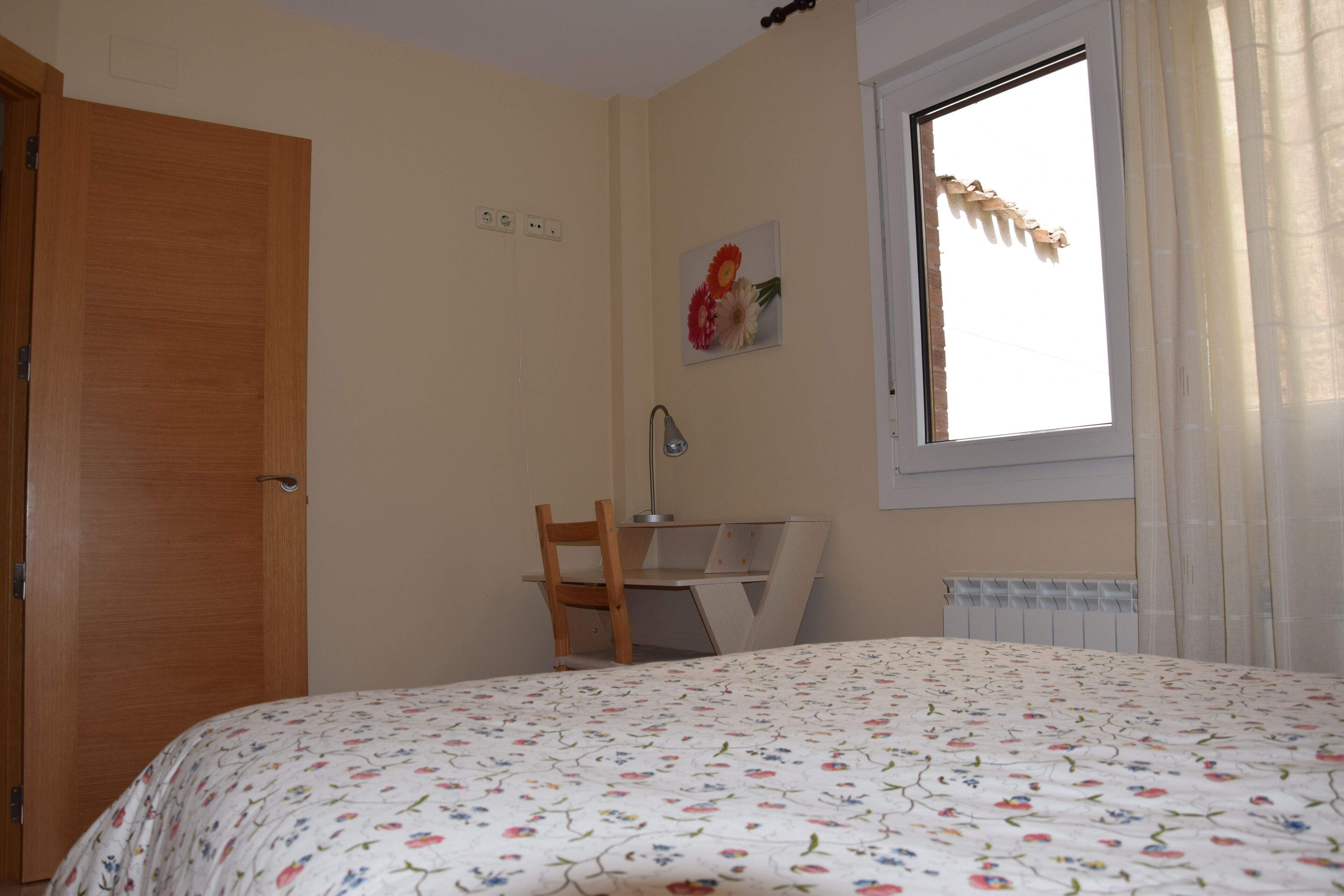 A bedroom with a bed, a wooden chair, a desk, a radiator, and a window with curtains.