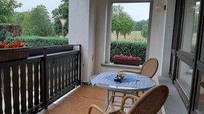 Ein Balkon mit einem runden Tisch, Korbstühlen, einem Blumentopf mit roten Blüten und Blick auf ein Feld und Bäume.