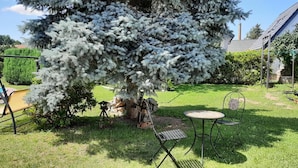 A backyard with a large tree, a yellow swing set, a metal table and chair, and a small garden area.