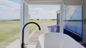 A modern bathroom with a large bathtub, a view of the ocean, and a wooden deck leading outside. - Wrights Bay House★Sea View★Private Beach★Robe (Mount Benson)
