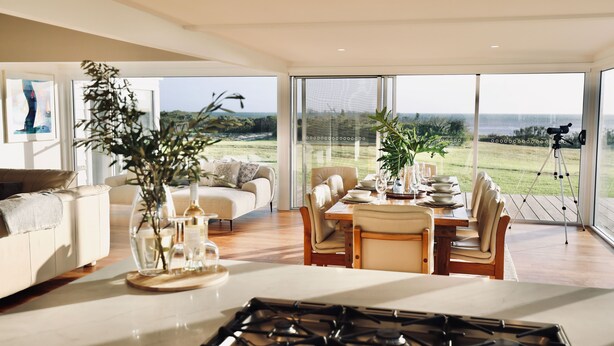 A modern living room with a large glass door leading to a balcony, a dining area with a table set for four, and a kitchen with a gas stove. - Wrights Bay House★Sea View★Private Beach★Robe (Mount Benson)
