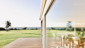 A modern outdoor deck with a wooden floor and a glass door leading to a dining area with white chairs and a table. - Wrights Bay House★Sea View★Private Beach★Robe (Mount Benson)