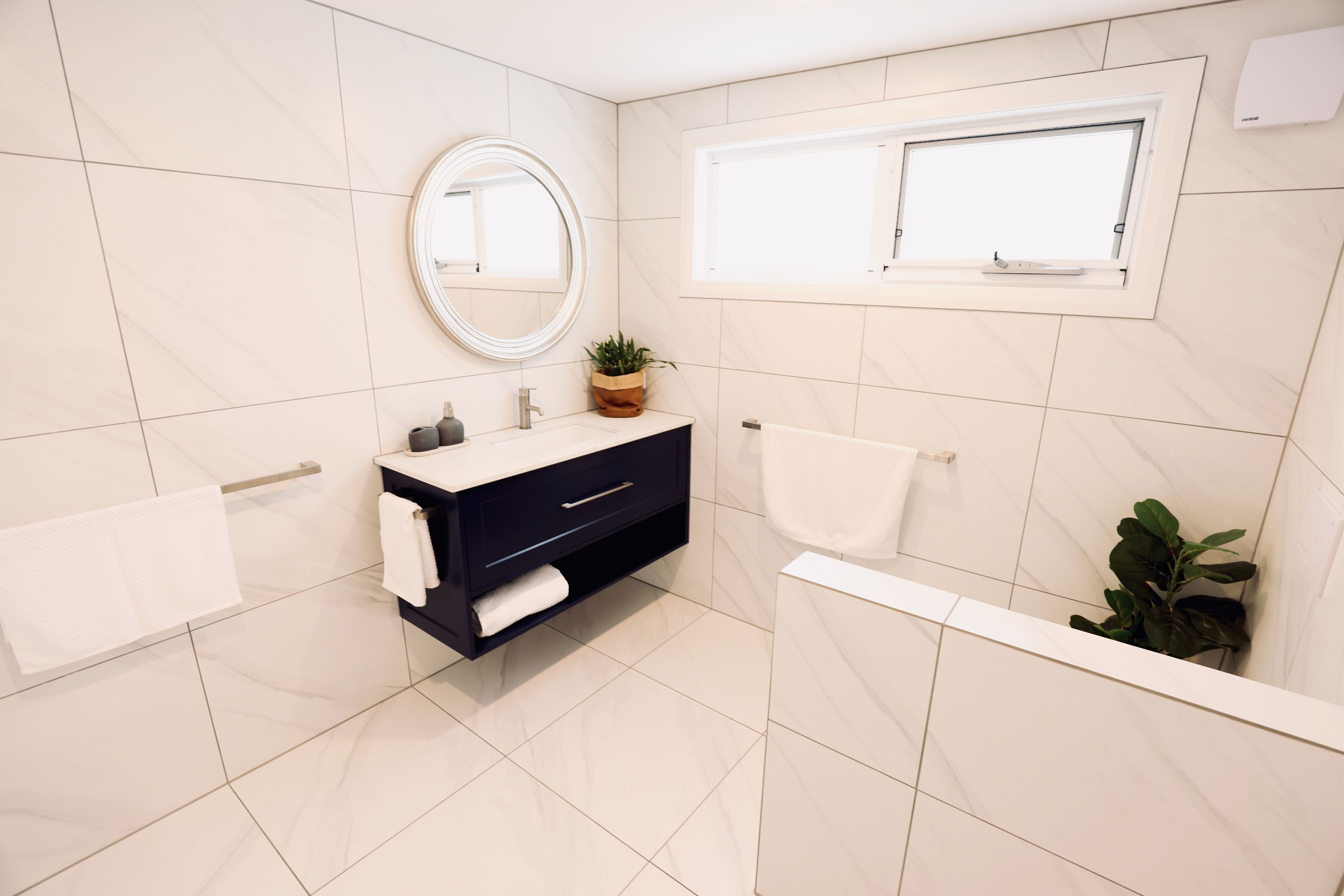 A modern bathroom with a dark vanity, a round mirror, a window, and a potted plant.