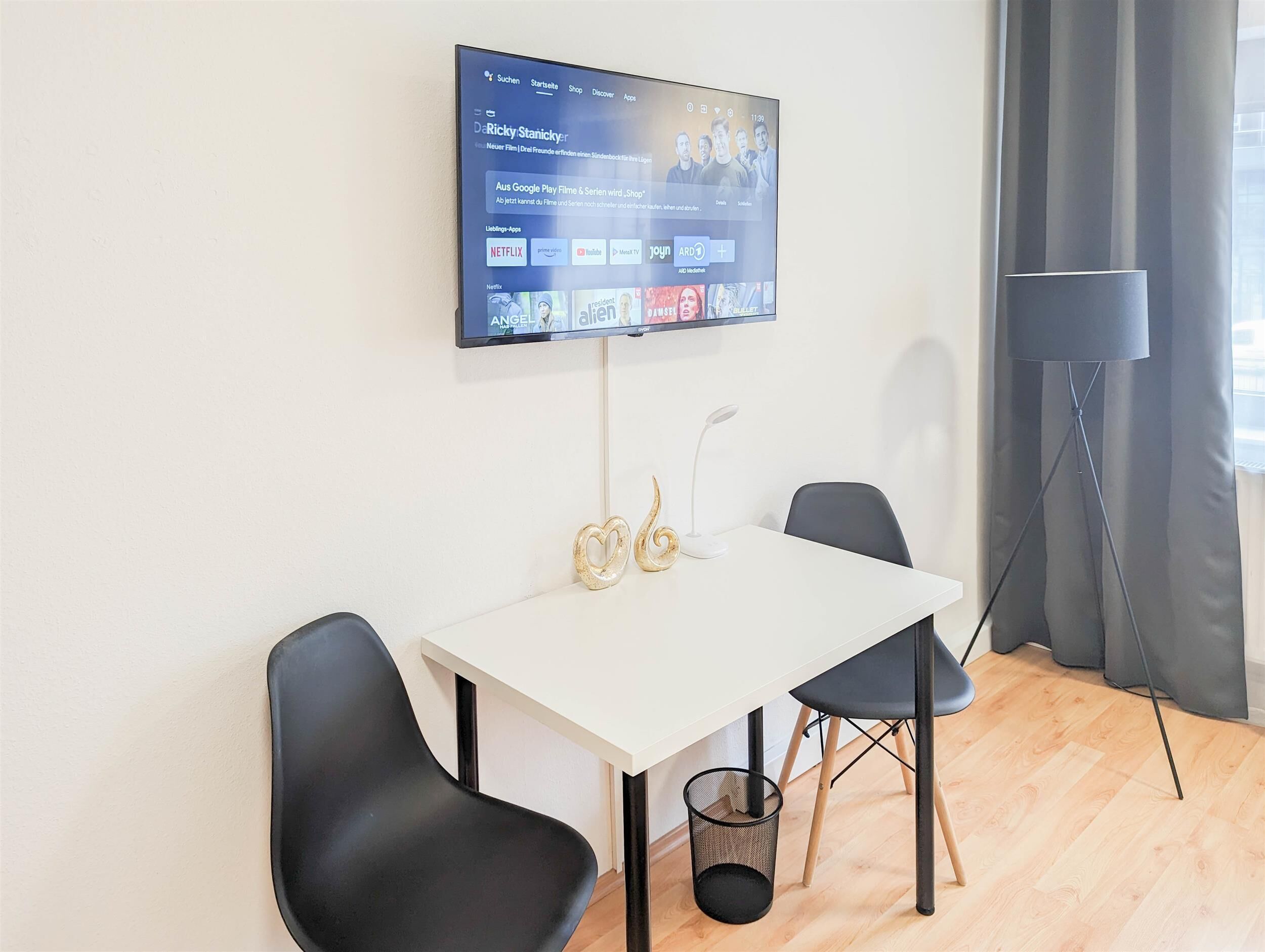 A modern room with a flat-screen TV mounted on the wall displaying a streaming service interface. There is a white table with two black chairs, a small decorative item, and a floor lamp.