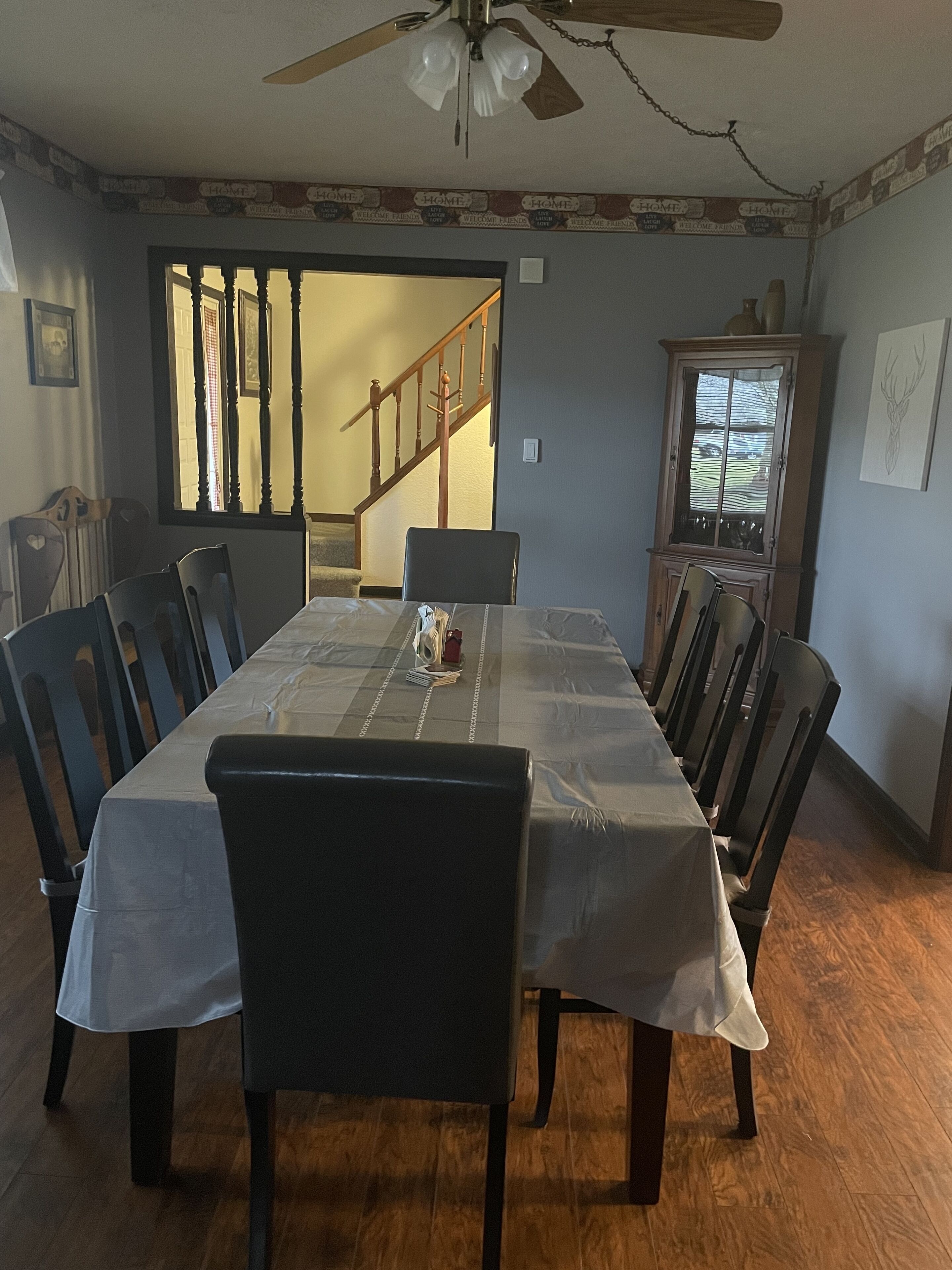 A dining room with a long table set for six, surrounded by chairs. A wooden cabinet with glass doors is against the wall, and a ceiling fan is visible above.