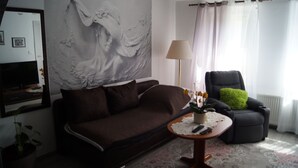 Uma sala de estar com um sofá marrom, uma mesa redonda pequena com um vaso, um travesseiro verde em uma cadeira preta e um mural grande na parede.