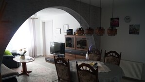 Uma área de refeições com uma mesa redonda e cadeiras, uma televisão ao fundo e uma cozinha com armários e luminárias pendentes.