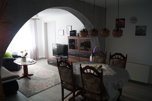 Uma área de refeições com uma mesa redonda e cadeiras, uma televisão ao fundo e uma cozinha com armários e luminárias pendentes.