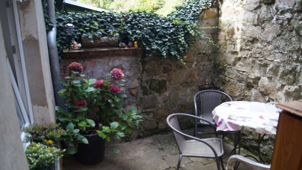 Uma pequena área de pátio com muro de pedra, planta em vaso com flores rosa, cadeira de metal e mesa com toalha branca.