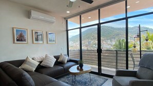 A modern living room with a dark brown sofa, a grey armchair, and a small round table. There are three framed pictures on the wall, and a ceiling fan is visible. - Gringo-Gulch, 3 bedroom w/view, pool/patio (Puerto Vallarta)