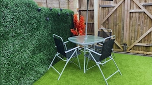 A patio with a round glass table and four chairs, a black umbrella, and a wooden fence in the background.