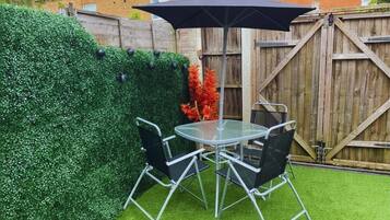 A patio with a round glass table and four chairs, a black umbrella, and a wooden fence in the background.