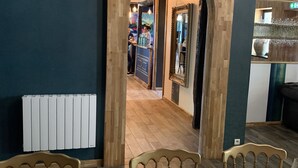 Un comedor con una mesa de madera dispuesta para una comida, un espejo y vista a otra habitación donde una persona trabaja en un mostrador.