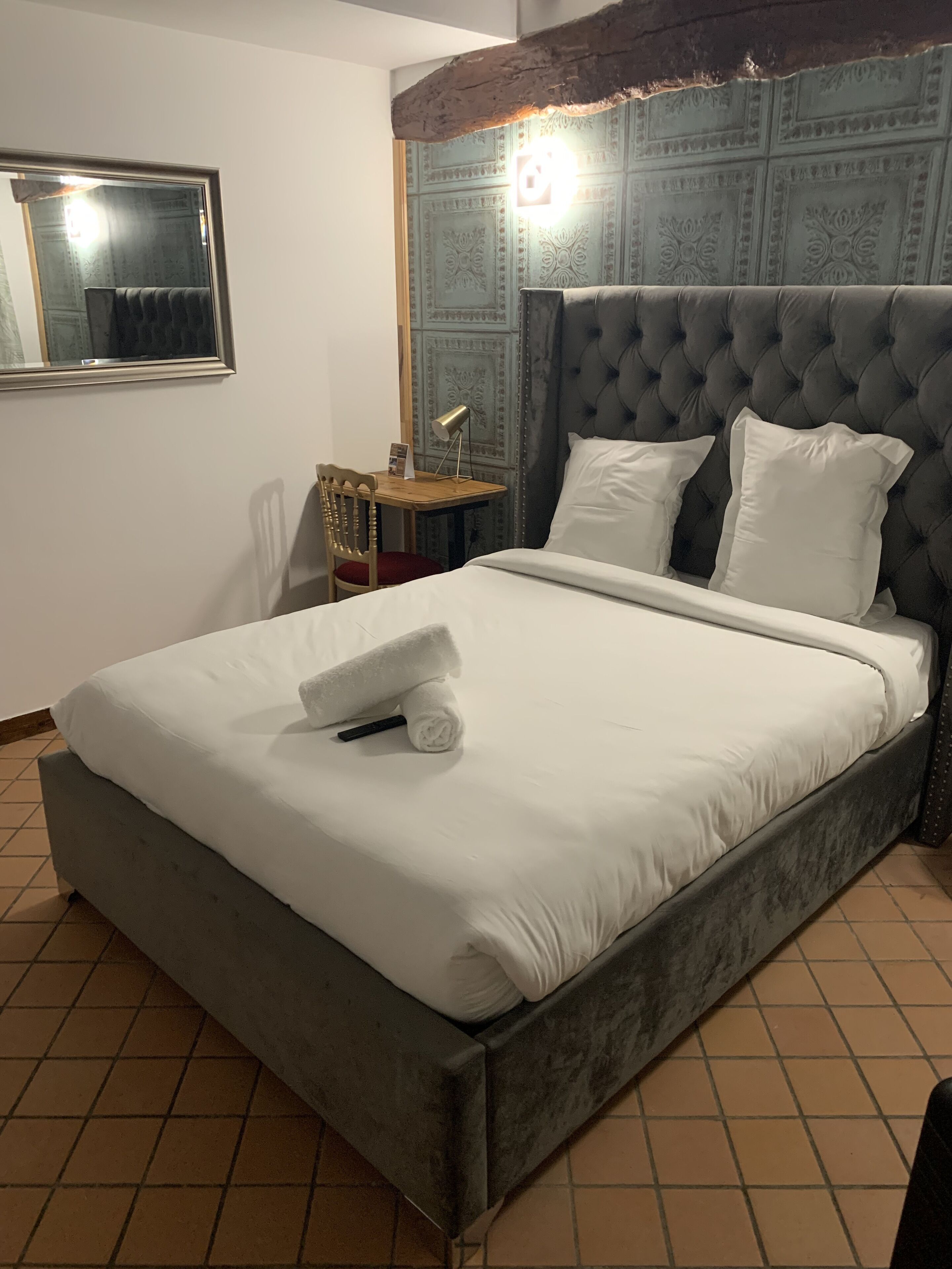 A neatly made bed with white linens, a grey upholstered headboard, and a bedside table with a lamp and a small item on it.