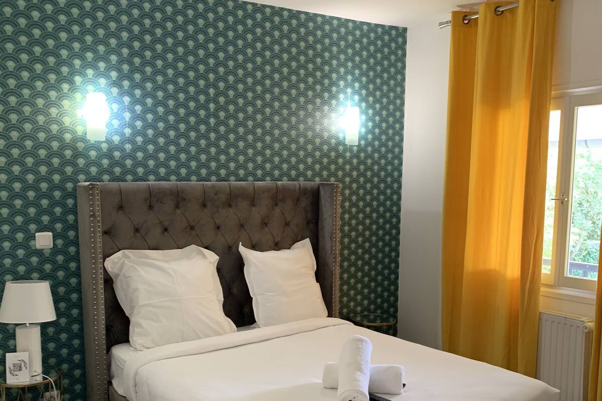 Una cama bien hecha con sábanas blancas y un cabecero marrón, una lámpara de noche y una ventana con cortinas amarillas.