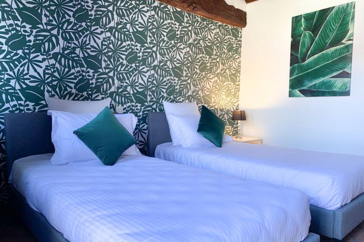 Una cama doble con ropa de cama blanca y almohadas verdes, una mesita de noche con lámpara, y un papel tapiz con estampado de hojas tropicales.