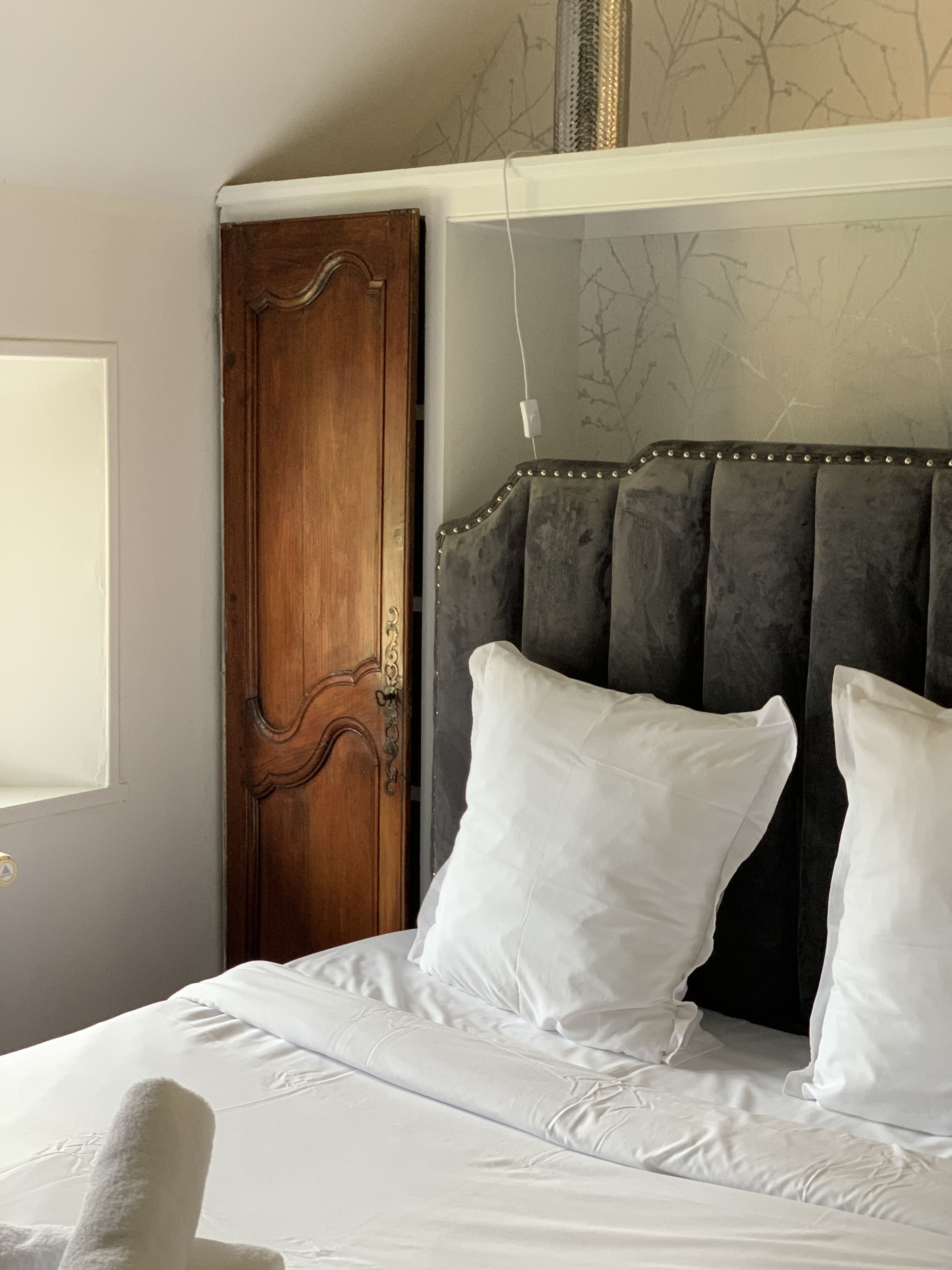 Un dormitorio con puerta de madera, cama con ropa blanca y cabecero tapizado oscuro.