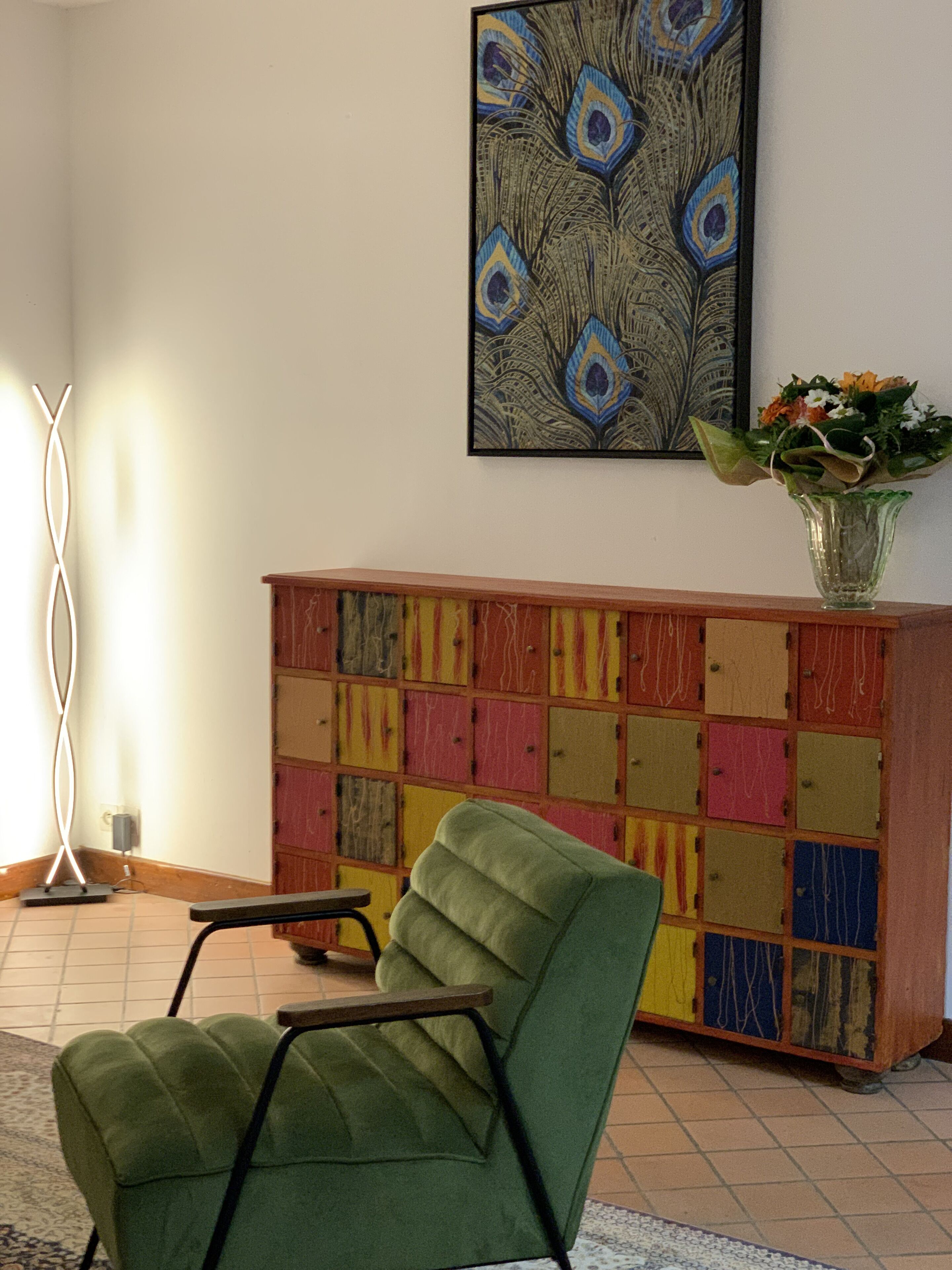 Una habitación con un sillón verde, un aparador colorido, un cuadro con motivos de pavo real y un jarrón con flores.