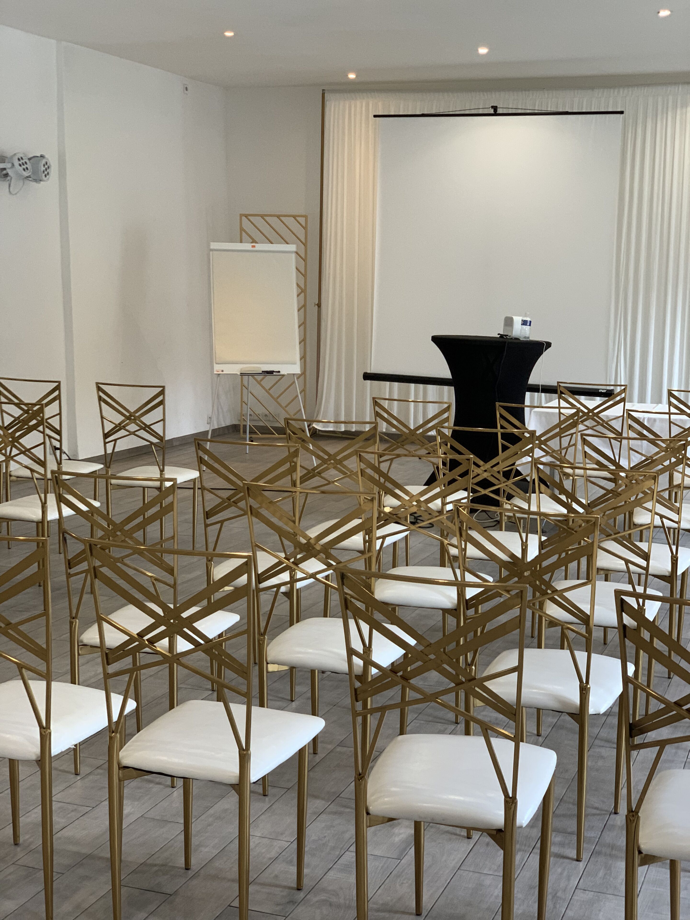 A conference room with rows of white chairs and a black table, white curtains, and a whiteboard on stands.