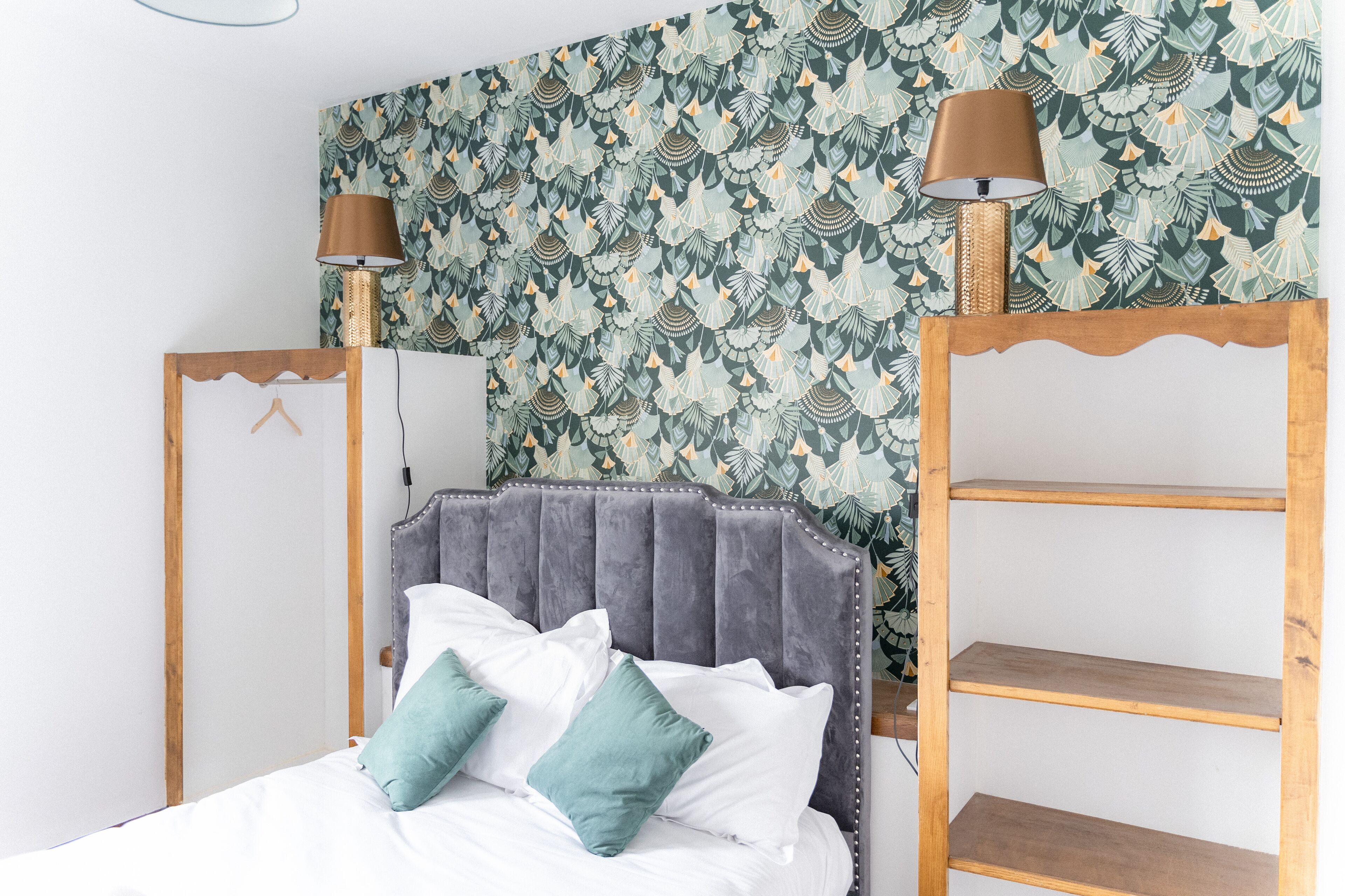 Un dormitorio con cabecera capitonada, cama con ropa de cama blanca y aguamarina, dos lámparas de pared y un papel tapiz estampado.