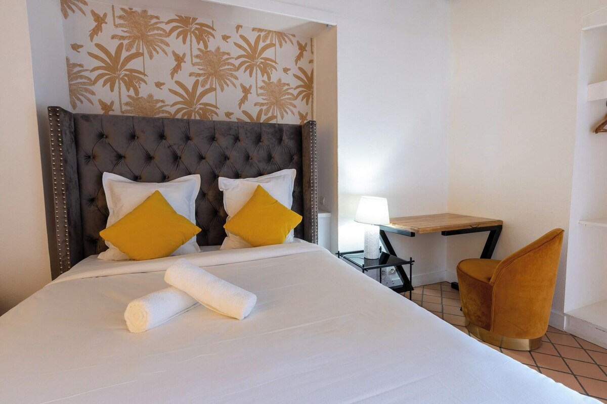 A neatly made bed with white linens and two yellow pillows, a tufted headboard, a small bedside table with a lamp, and a chair with a wooden frame.