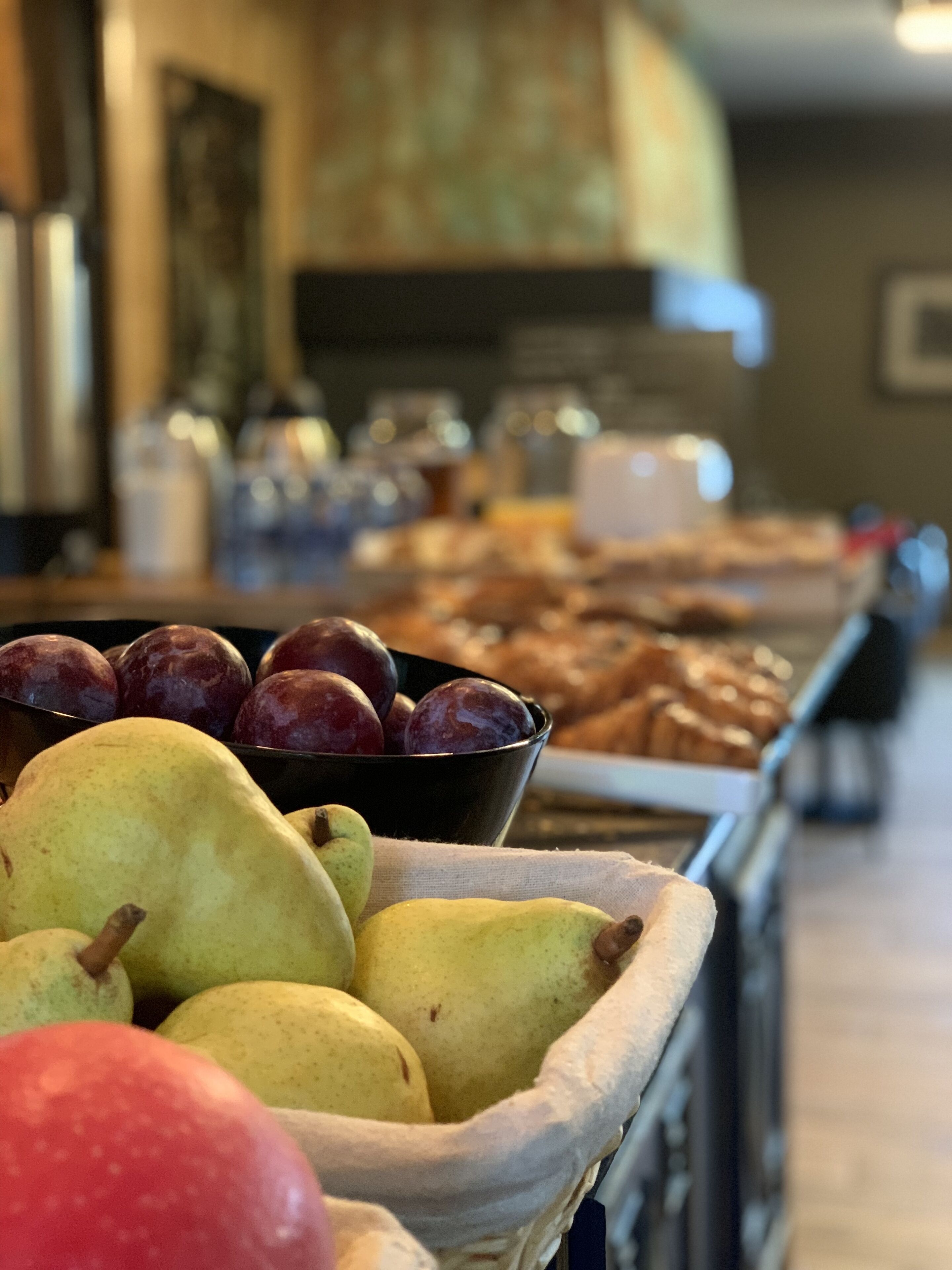 Una cesta de frutas con manzanas y ciruelas, una cafetera y pasteles al fondo.
