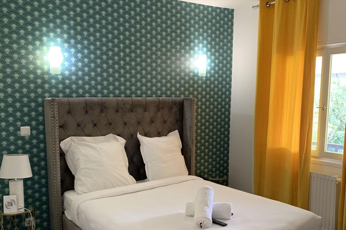 A neatly made bed in a room with a patterned wallpaper, a bedside lamp, and a window with yellow curtains.