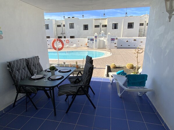 A balcony with a dining table set for two, a pool in the background, and a view of a building complex.