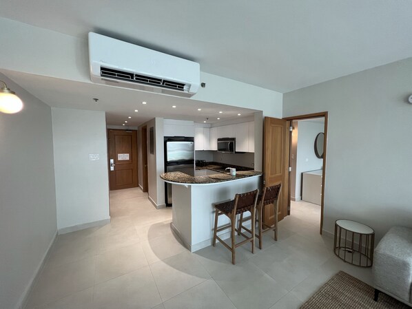 Cocina compacta con una isla central, un refrigerador y un microondas. Una puerta de madera conduce a otra habitación.