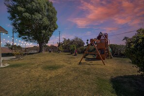A backyard with a swing set, a tree, and a house in the background. - Charming 5BRS/3BTHS - DISNEY - CONVENTION - PLAYGROUND - SOCAL FUN! (Anaheim)