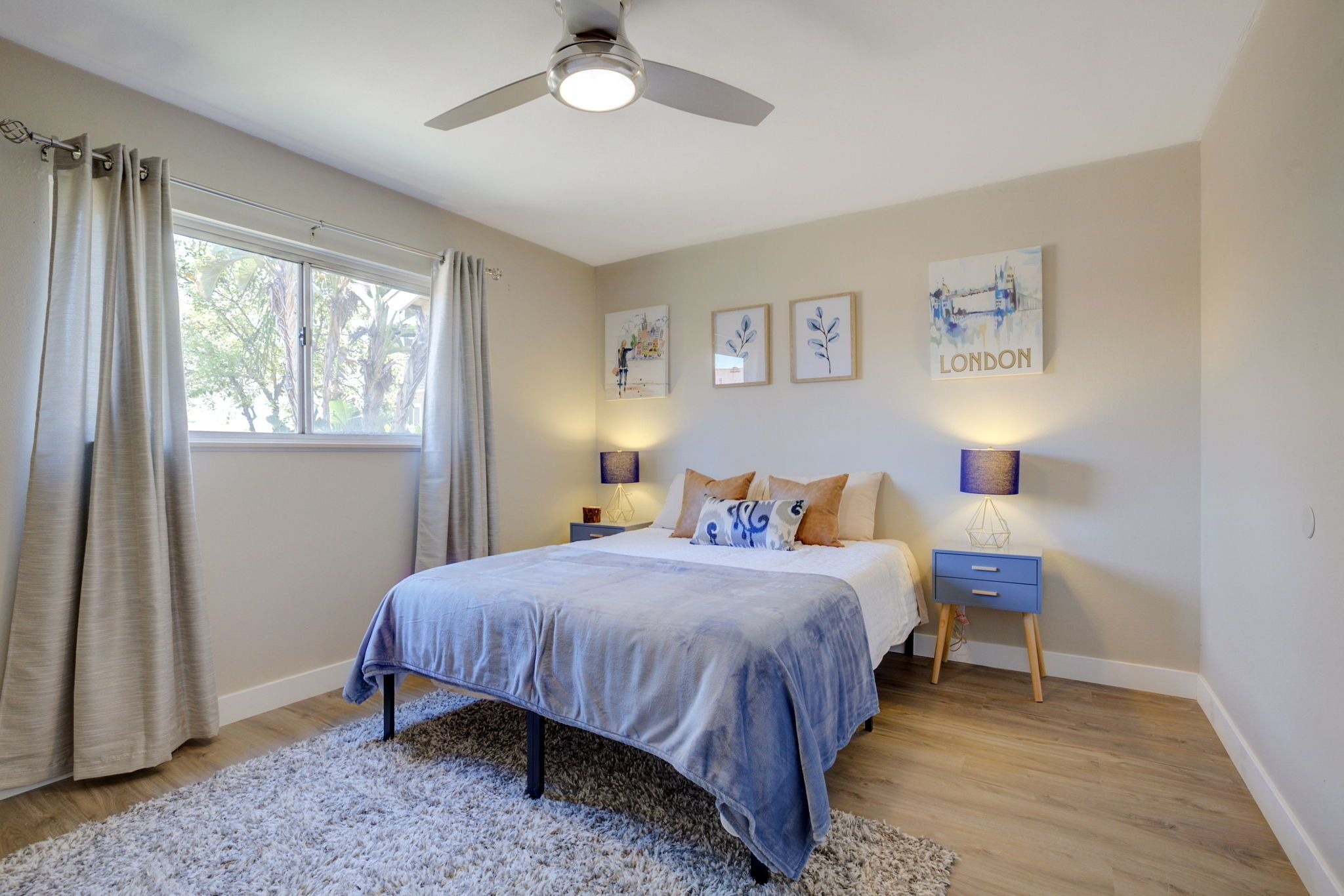 Une chambre à coucher comprenant un lit, un ventilateur de plafond, une fenêtre avec des rideaux, des œuvres d’art murales et des lampes de chevet.