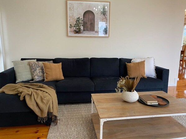 Un sofá oscuro con cojines estampados, una mesa de centro de madera con objetos decorativos y un cuadro en la pared.