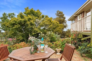 A wooden table with a bottle and glasses, surrounded by lush greenery and flowering plants.