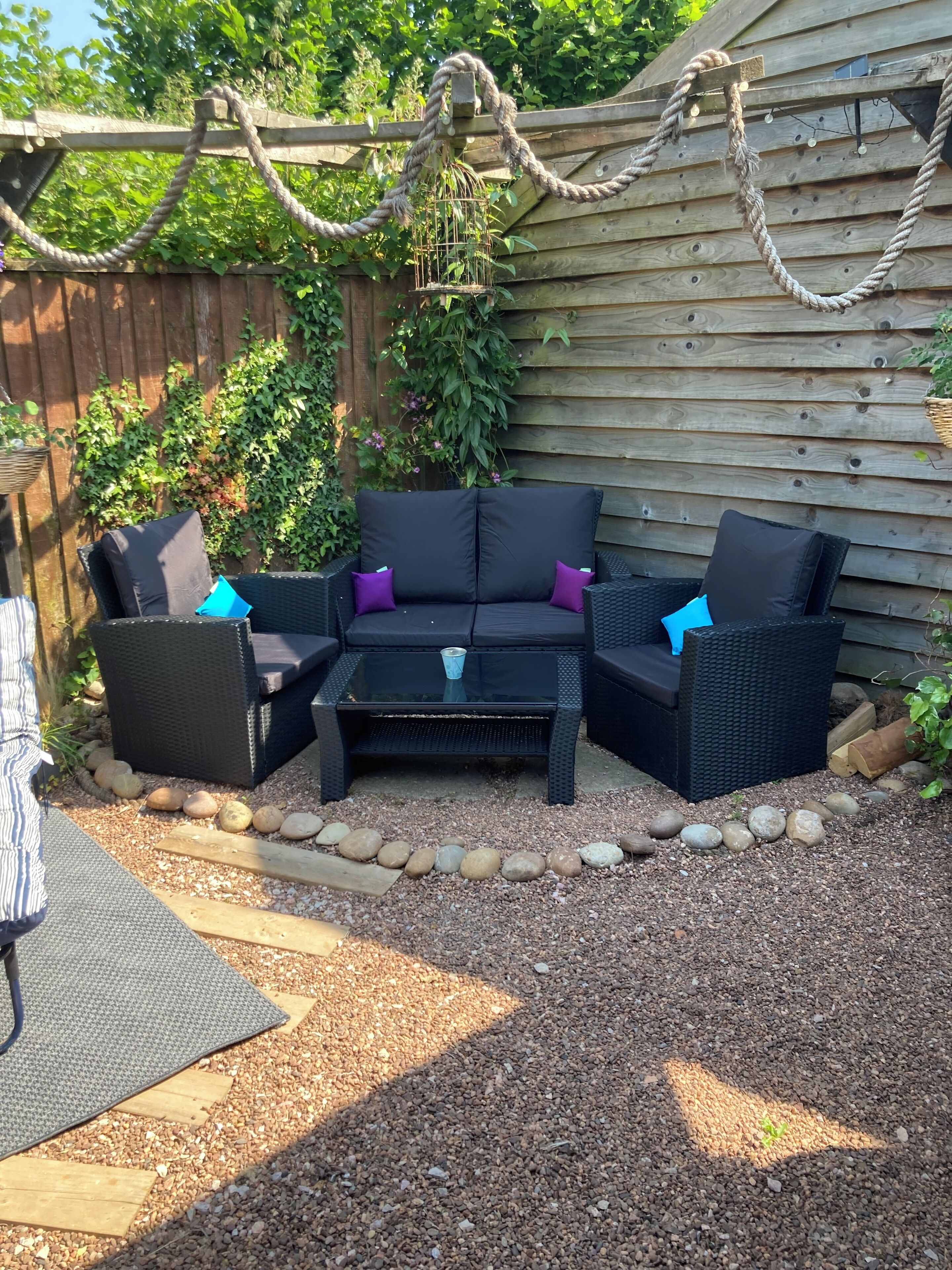 A patio area with a black wicker sofa set, a glass coffee table, and a small table with a blue cup. There is a wooden fence in the background and a garden with various plants.