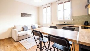 Ein kompakter Wohnraum mit Sofa, Esstisch und Stühlen, einer Küchenzeile und großen Fenstern.