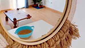 Ein runder Spiegel mit Kordelrahmen, der einen Raum mit einem Holztisch, einer blauen Schüssel, einem an der Wand hängenden Fernseher und Zimmerpflanzen reflektiert.