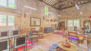 A spacious room with a high ceiling, wooden beams, and large windows. There are several wooden chairs and a sofa arranged around a central area with a patterned rug. - DOMAINE DU MOULIN D'ANDRAUT Bordeaux-horses-golf-St Emilion (Saint-Sulpice-et-Cameyrac)