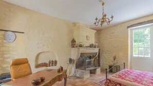 A room with a wooden table, a chair, a fireplace, a bed with a floral bedspread, and a window with a view of greenery. - DOMAINE DU MOULIN D'ANDRAUT Bordeaux-horses-golf-St Emilion (Saint-Sulpice-et-Cameyrac)