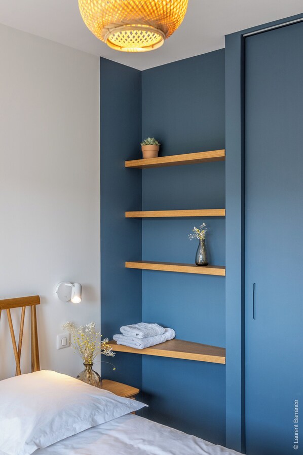 A bedroom with a bed, a nightstand, a wall-mounted shelf with a potted plant, a vase with flowers, and a towel rack.