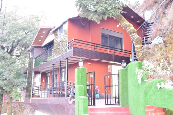 Un immeuble de deux étages avec une façade rouge, un balcon et un escalier menant à l’entrée.